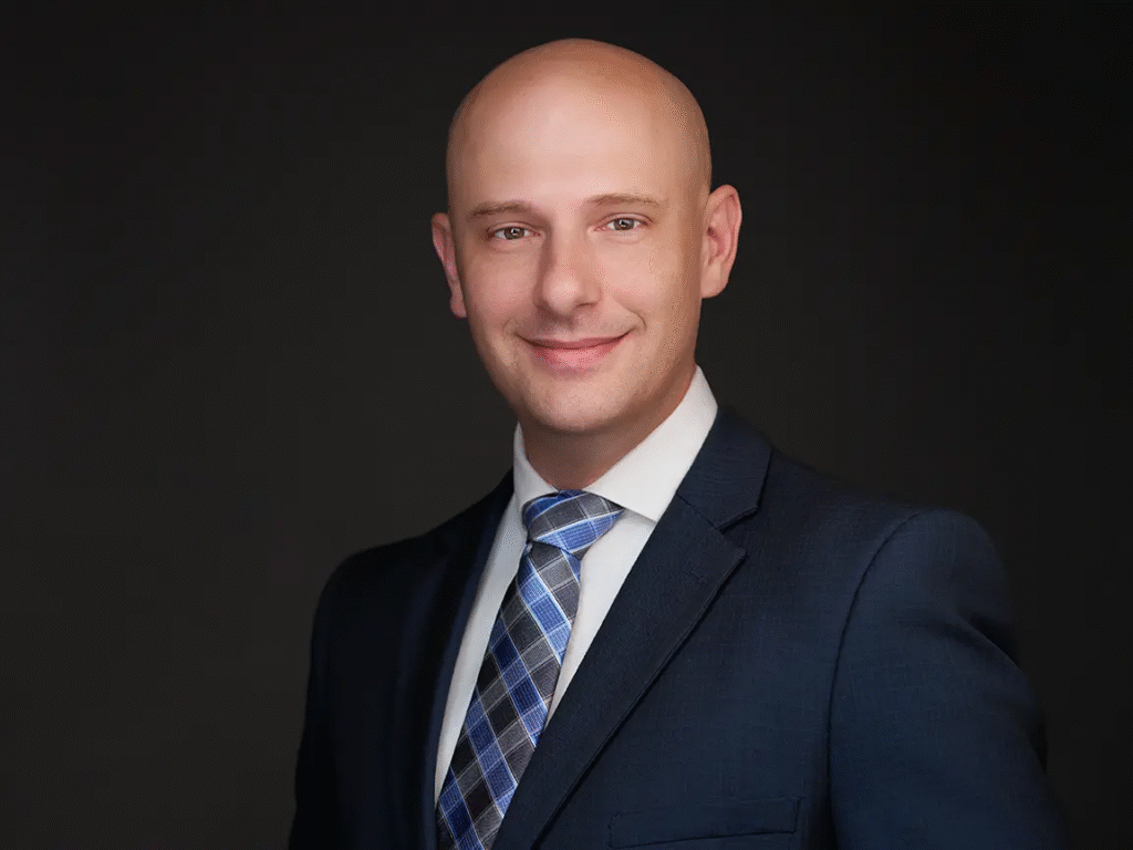 Professional headshot of a man in a navy suit and blue patterned tie.
