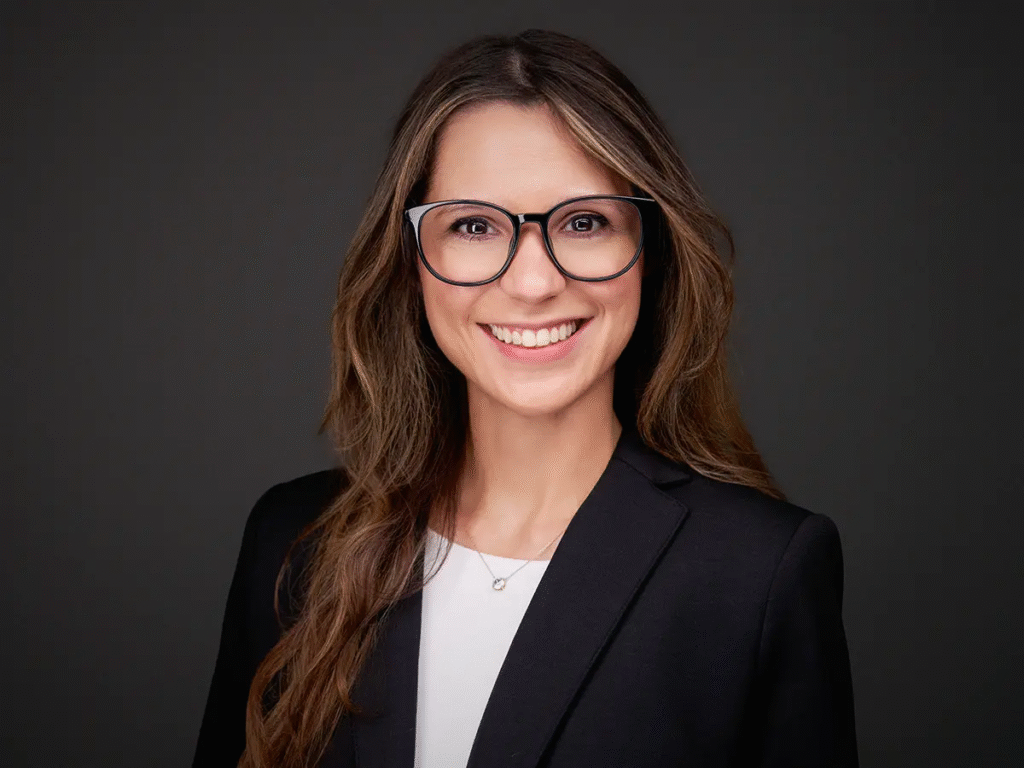 Professional headshot of a woman wearing glasses and a black blazer, smiling against a dark background.