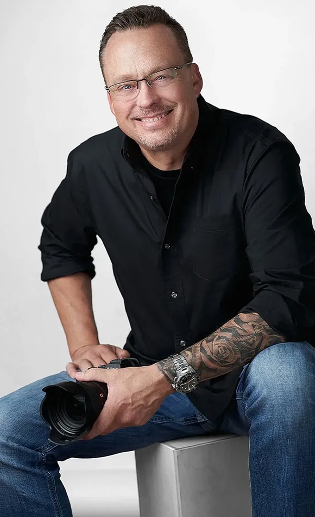 Man with glasses, wearing a black shirt and jeans, sits and smiles at the camera, holding his own camera. His forearm tattoo adds character to this relaxed yet professional headshot. Perfect example of Men's Headshots in San Antonio.