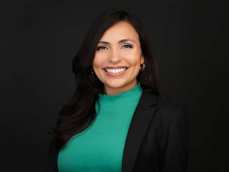 Top 10 Ways Business Headshots Improve Your Personal Brand 29 Smiling person in a teal top and black blazer against a dark background.