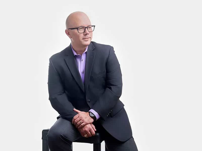 Top 10 Ways Business Headshots Improve Your Personal Brand 30 Person wearing glasses in a dark blazer and purple shirt, seated on a stool against a white background.