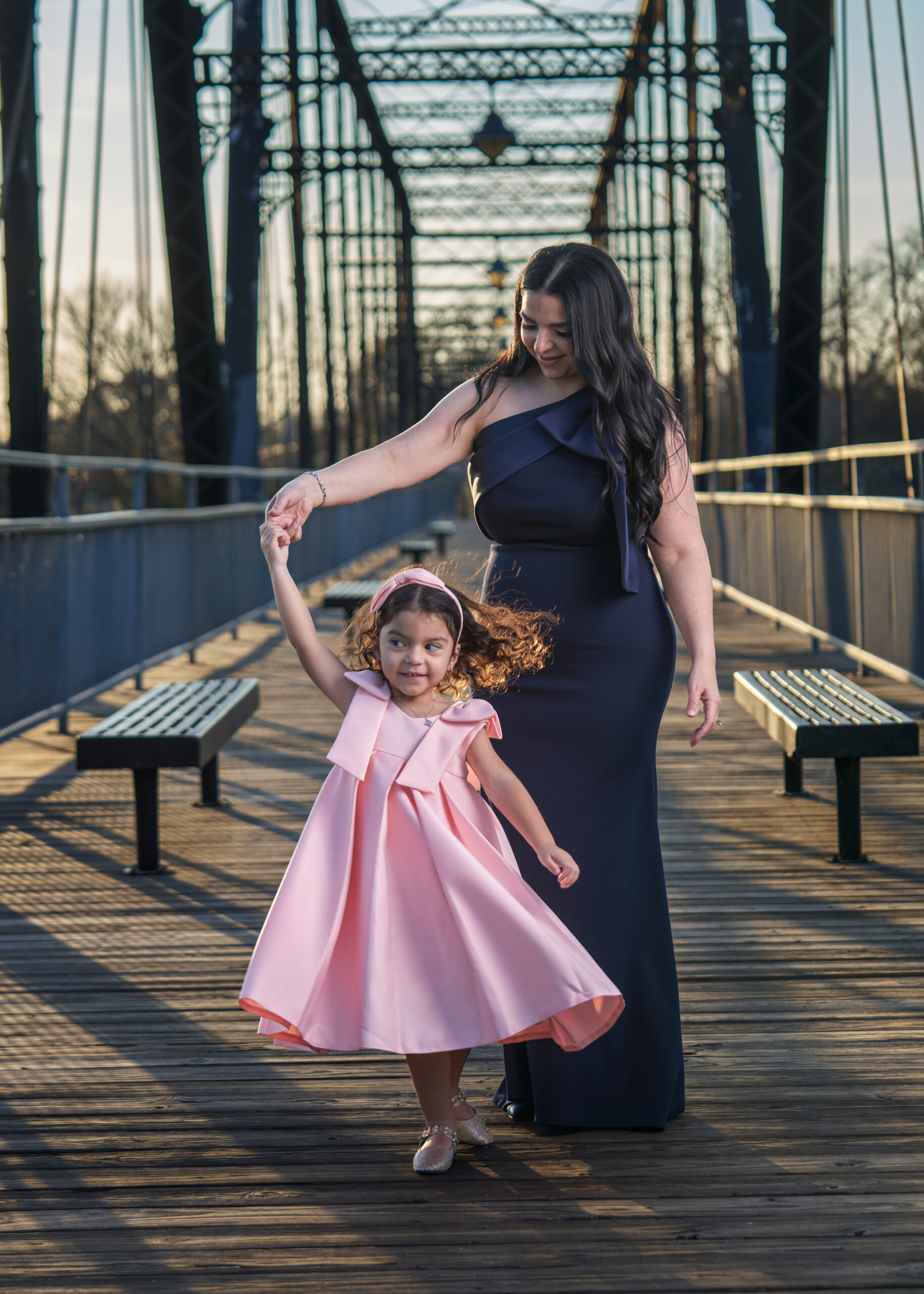 A woman in a long black dress twirls a young girl in a pink dress on a wooden bridge with metal railings, benches, and a framework structure—an enchanting scene perfect for personal headshots in a natural outdoor setting.