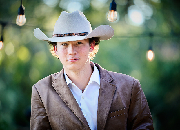 A young man in a tan cowboy hat and brown jacket stands outdoors with hanging lightbulbs and greenery in the background, creating a unique personal headshot setting.