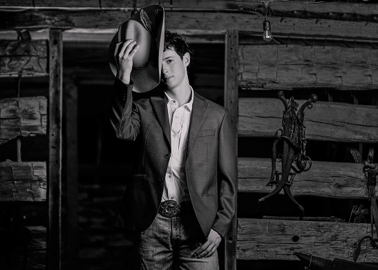 A young man in a suit jacket and jeans stands in front of a rustic wooden wall, holding a cowboy hat partly over his face—an authentic take on Men's Headshots San Antonio style.