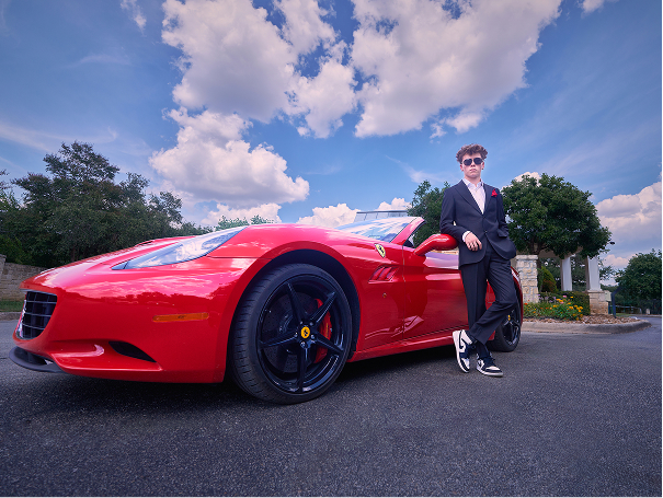 A man in a black suit and sunglasses leans against a red Ferrari sports car parked outdoors under a partly cloudy sky, showcasing confidence and style perfect for men's headshots in San Antonio.