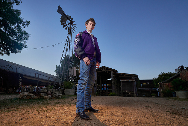 A young man in a purple letterman jacket and jeans stands outdoors on a dirt path at dusk, with buildings and a windmill in the background—an ideal scene for a personal headshot.