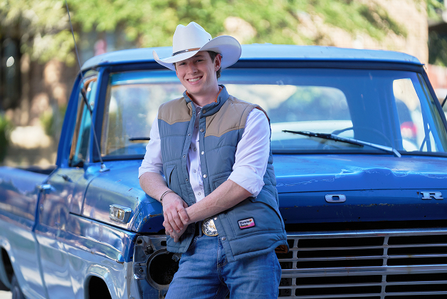 A person in a white cowboy hat and vest leans against the front of a vintage blue Ford truck, smiling at the camera—capturing a relaxed take on men’s headshots with a touch of professional style.