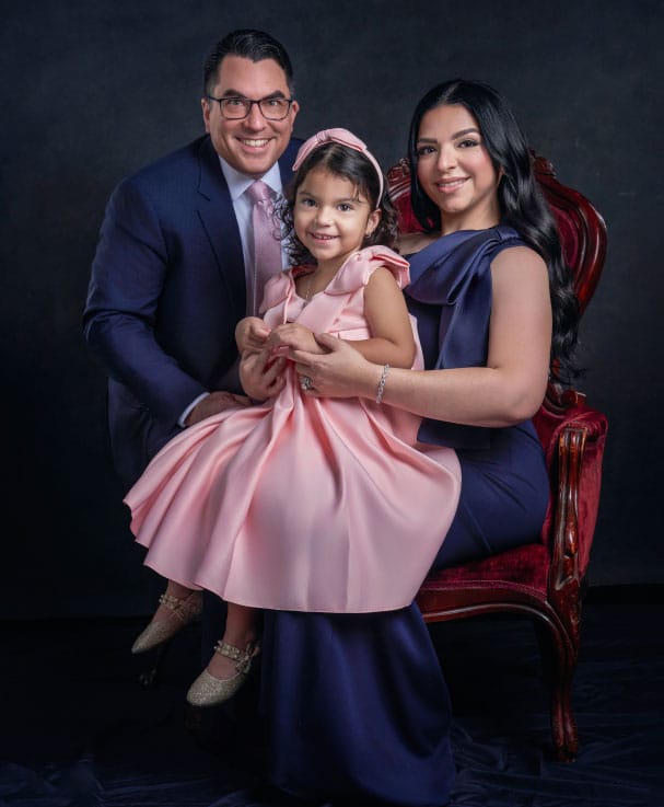 A man in a suit, a woman in a dark gown, and a young girl in a pink dress pose together for a formal family portrait against a dark background—an elegant image perfect for personal headshot inspiration or legal headshots.