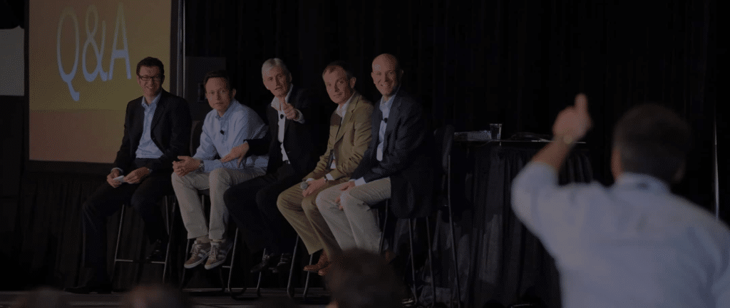 How Conference Photography Supports Your Marketing Team 28 Five men sit on a stage during a Q&A panel while an audience member in the foreground raises a hand to ask a question.