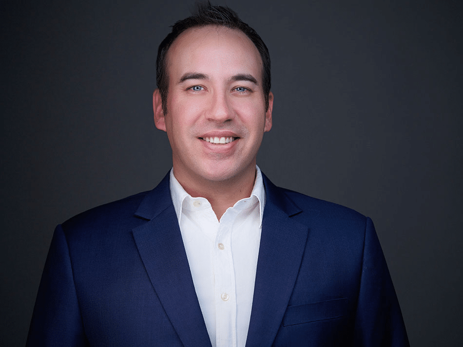 What Attorneys Should Wear For Headshots 28 Man in a dark blue suit and white shirt smiling in front of a plain dark background.