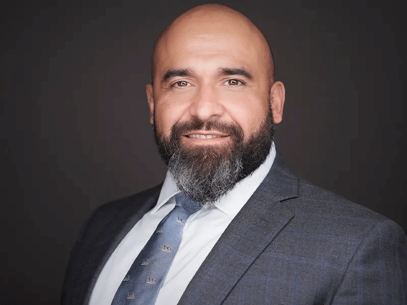 What Attorneys Should Wear For Headshots 29 A man with a shaved head and full beard wearing a grey suit, white shirt, and blue tie poses against a plain dark background.