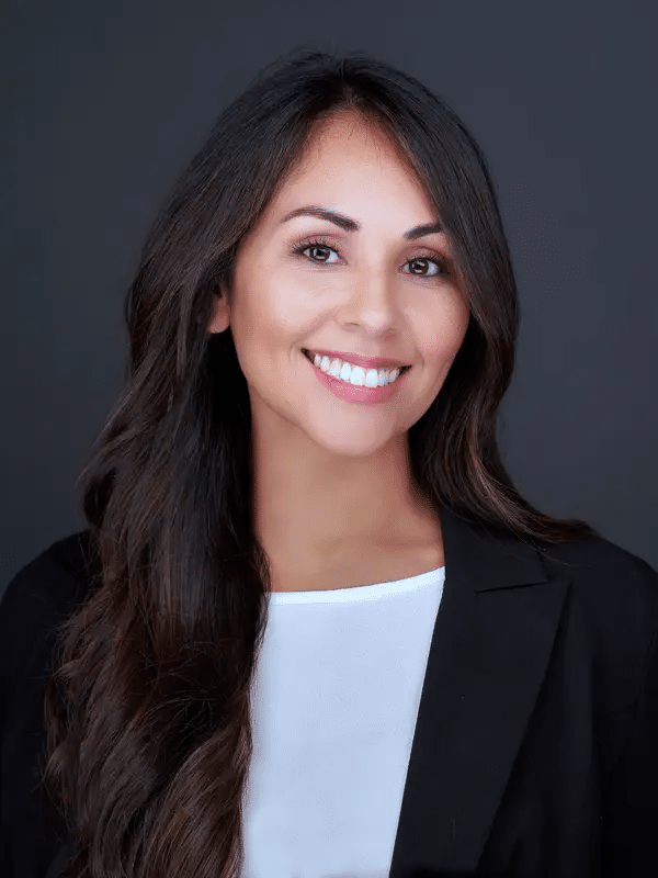 What Attorneys Should Wear For Headshots 30 Woman with long dark hair wearing a black blazer and white blouse, smiling at the camera against a plain dark background.