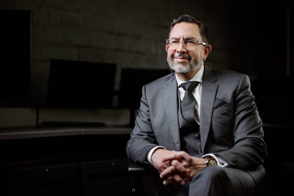 A man in a gray suit and glasses sits in an office chair, smiling with his hands folded, in front of a dark background with computer monitors—capturing the polished professionalism often sought by Matt Roberts, a photographer based in San Antonio known for expert headshots for women and men alike.