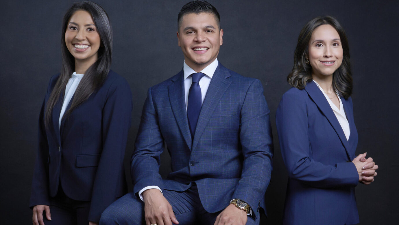 Professional portrait of three business professionals in formal navy blue business attire posing together against a dark background.