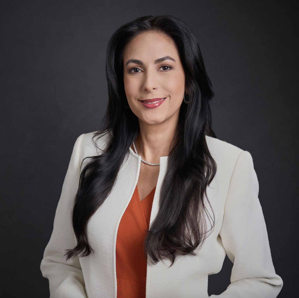 Professional business headshot of a woman in a white blazer and orange blouse against a dark studio background.