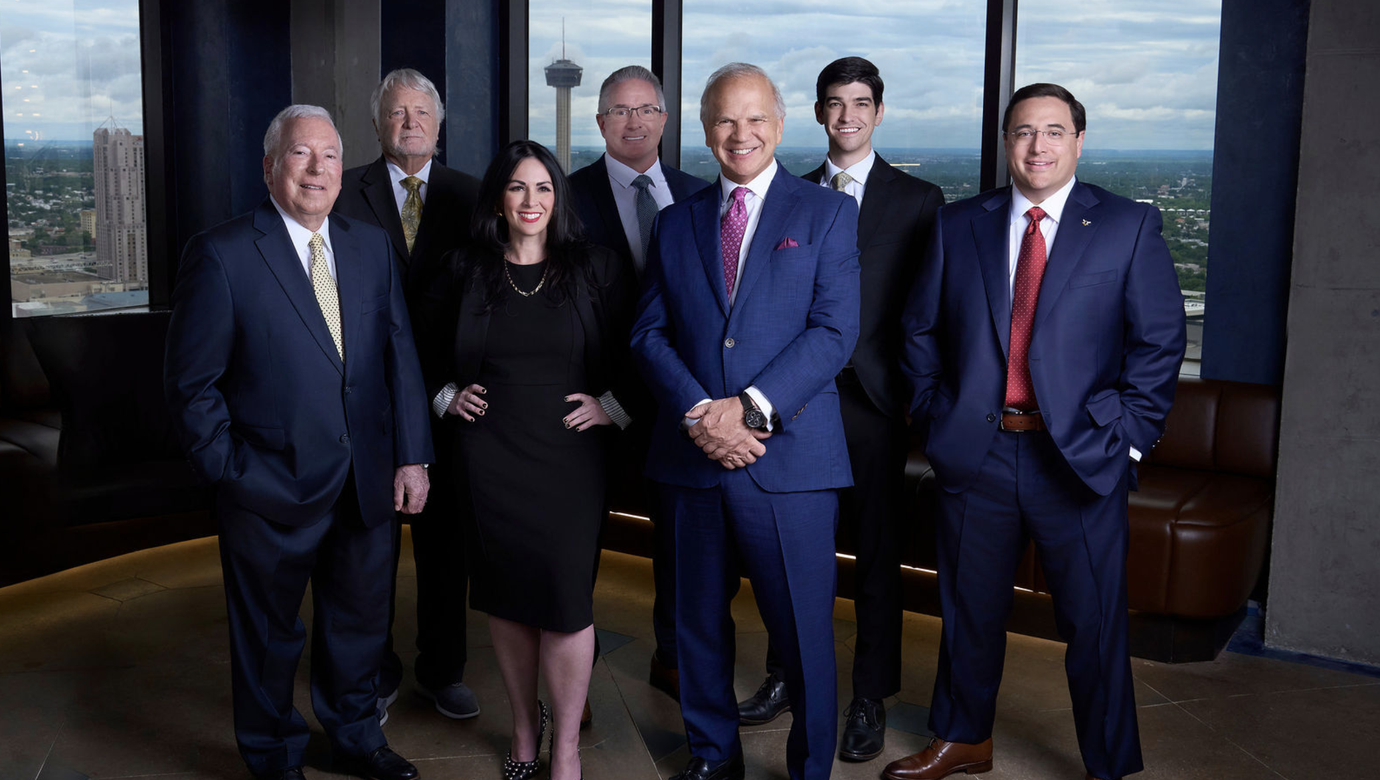 Professional corporate team portrait of seven business professionals in a high-rise setting with the San Antonio skyline visible through the windows.